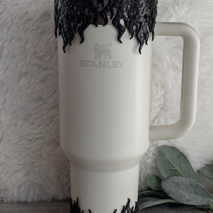 May include: White Stanley travel mug with a black flame design around the top and bottom. The mug has a handle and a straw.