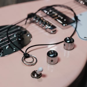 May include: Close-up of a light pink electric guitar. The guitar features chrome hardware, including the bridge, pickups, and control knobs. Black strings and wiring are visible against the pink body. A white pickguard is also present.