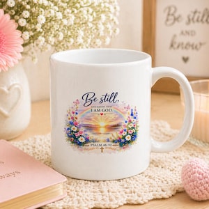 Op de afbeelding: Witte keramische mok met de tekst "Be still, and know that I AM GOD" omgeven door een bloemen- en regenboogontwerp. Een roze boek met "Jesus Always" staat naast de mok. Een kaars en een ingelijste print staan op de achtergrond.