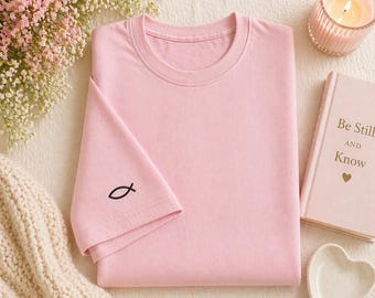 May include: A light pink t-shirt with a black fish symbol embroidered on the sleeve. The shirt is folded neatly, with a book, candle, and flowers in the background. The book reads "Be Still and Know".