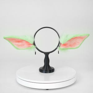 May include: A pair of plush, light green and pink costume ears attached to a black headband. The ears are fluffy with pink inner ear details. The headband is supported by a black stand on a white surface.