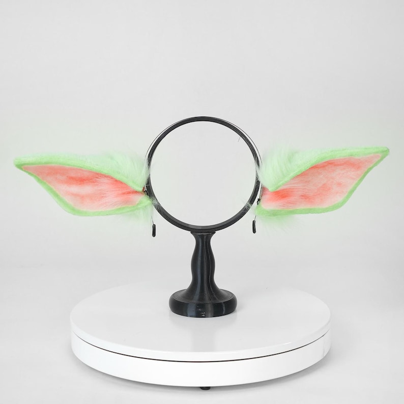 May include: A pair of plush, light green and pink costume ears attached to a black headband. The ears are fluffy with pink inner ear details. The headband is supported by a black stand on a white surface.