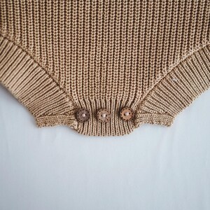 Puede incluir: Primer plano de un pelele tejido de color beige con tres botones de madera en la parte inferior. El pelele tiene una textura acanalada y un color neutro suave. Los botones son redondos y de color marr&oacute;n claro.