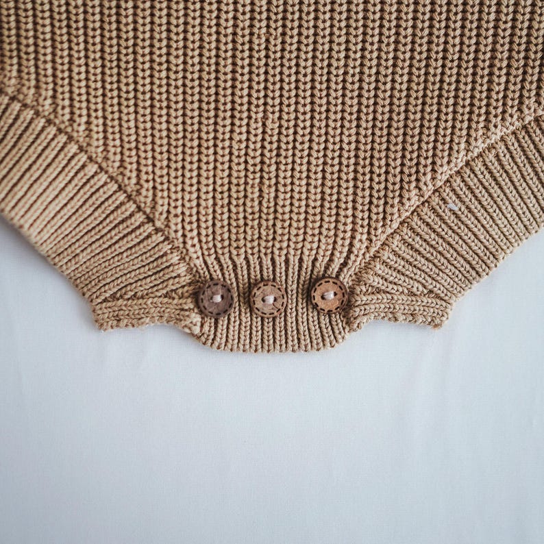 Puede incluir: Primer plano de un pelele tejido de color beige con tres botones de madera en la parte inferior. El pelele tiene una textura acanalada y un color neutro suave. Los botones son redondos y de color marr&oacute;n claro.