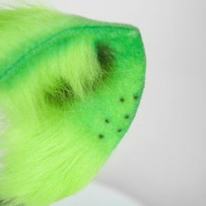 May include: A bright neon green faux fur ear accessory with a soft, plush texture. The ear features a pointed shape with a darker green inner section and a series of small, evenly spaced black dots near the outer edge of the fabric surface.
