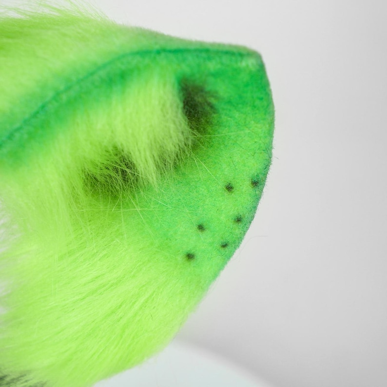 May include: A bright neon green faux fur ear accessory with a soft, plush texture. The ear features a pointed shape with a darker green inner section and a series of small, evenly spaced black dots near the outer edge of the fabric surface.