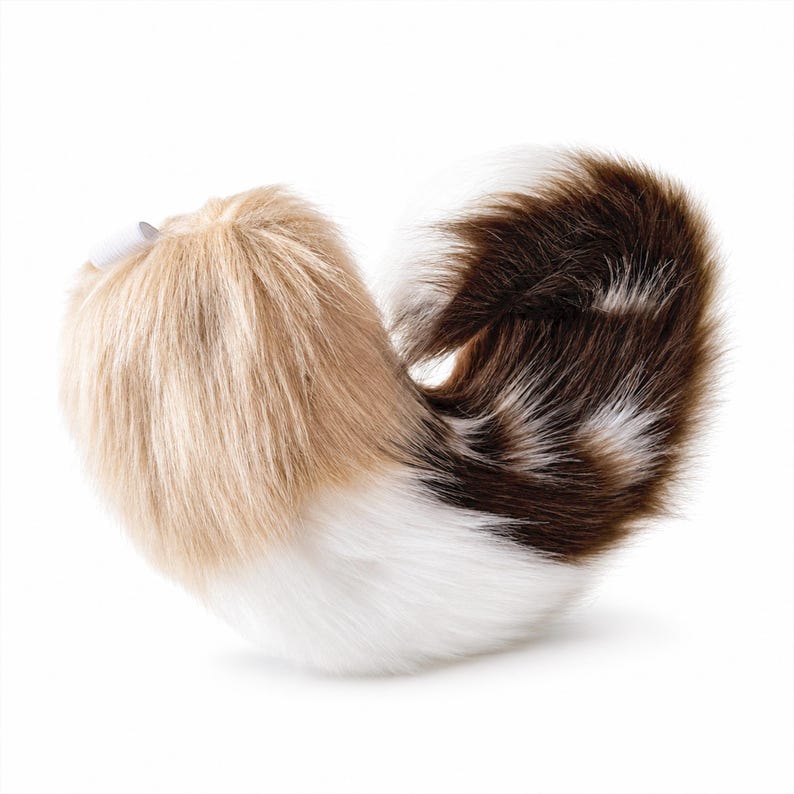 May include: A curved, plush faux fur tail accessory featuring a tri-color pattern of light tan, white, and dark brown. The texture is long and soft, with a small white fabric loop attached at the top end for secure fastening or attachment.