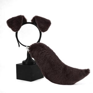 May include: A dark brown plush animal ear headband and a matching faux fur tail. The headband features two soft, rounded ears attached to a thin black frame, while the curved, tapered tail includes a small white attachment clip at the base.