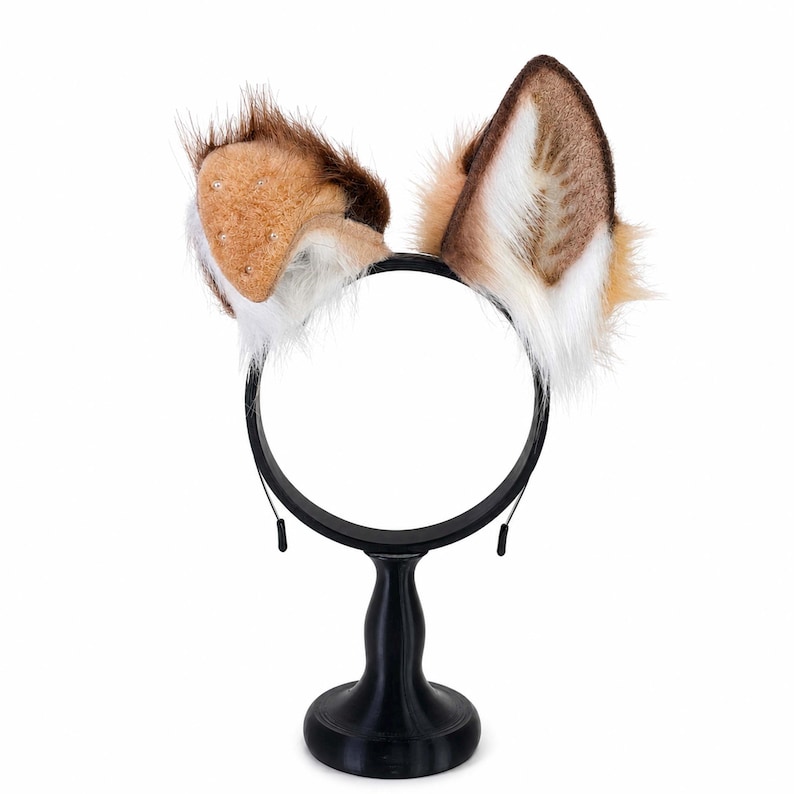 May include: A black headband featuring two faux fur animal ears. The ears are colored in shades of brown, tan, and white, with long, textured fur accents. The left ear is rounded and the right ear is pointed, both attached to a thin black band.