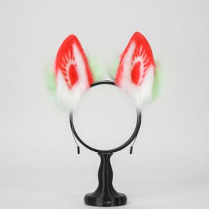 May include: A black headband featuring two upright faux fur ears. The ears have a white base, a bright red center with a radiating pattern, and light green fur accents along the outer edges. The headband is mounted on a black circular stand.