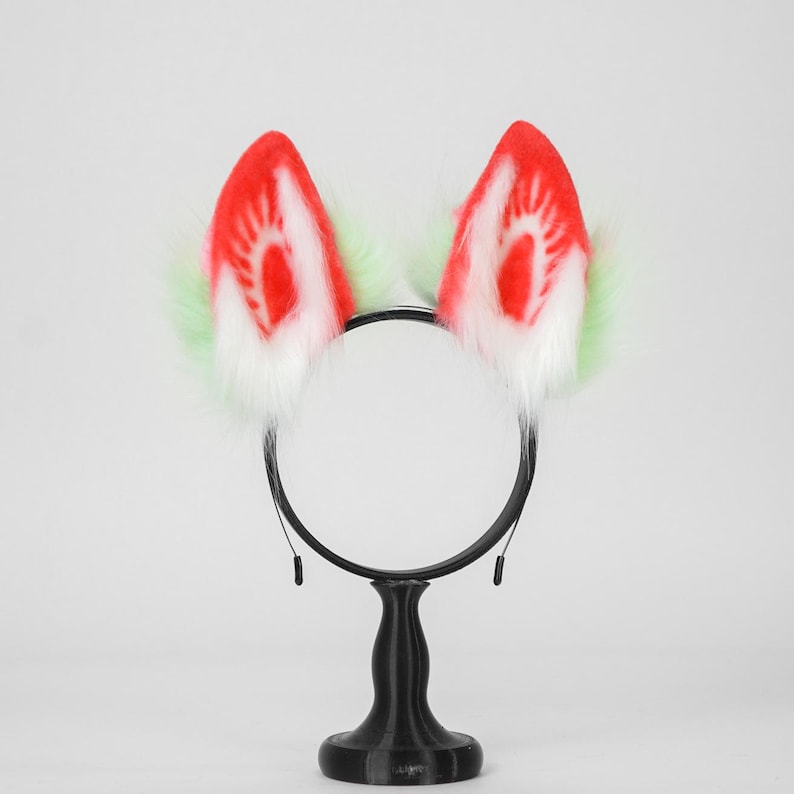 May include: A black headband featuring two upright faux fur ears. The ears have a white base, a bright red center with a radiating pattern, and light green fur accents along the outer edges. The headband is mounted on a black circular stand.