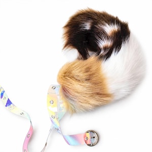 May include: A faux fur tail accessory featuring a tri-color pattern of white, brown, and tan long fibers. The tail is attached to a long, iridescent, holographic-finish strap made of a smooth material, featuring metal eyelets and a silver buckle.