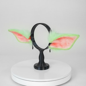 May include: A black headband with attached green and pink faux fur ears. The ears are shaped like stylized animal ears, with a fluffy texture and a contrasting color scheme. The headband is displayed on a white surface.