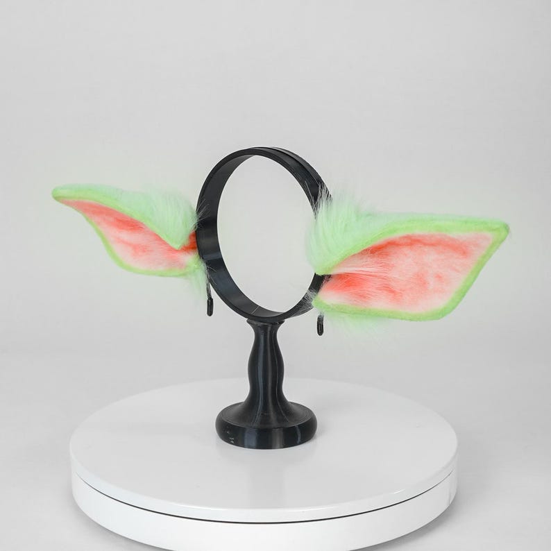 May include: A black headband with attached green and pink faux fur ears. The ears are shaped like stylized animal ears, with a fluffy texture and a contrasting color scheme. The headband is displayed on a white surface.