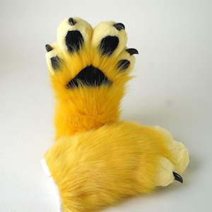 May include: A pair of bright yellow faux fur paw gloves featuring four padded fingers and a palm pad in black. Each finger is tipped with a curved, pointed black claw. The plush, long-pile yellow fur covers the entire exterior of the hand-shaped gloves.