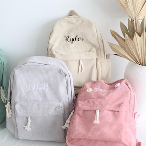 May include: A collection of corduroy backpacks in various colors: light beige, gray, and pink. Each backpack features a front pocket, a top handle, and the name of the owner embroidered on the front. The backpacks have white rope-like drawstrings.