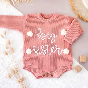 May include: A pink knitted baby romper with long sleeves. The words "big sister" are embroidered in white script across the front, with small white cloud-shaped accents. The romper has three wooden buttons at the bottom.