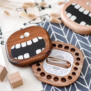 May include: Wooden tooth keepsake boxes with cartoon faces. One box is open, revealing a chart for baby teeth. A small glass vial is inside. The boxes are brown with white and black details. The background includes a white and blue patterned cloth.