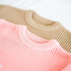 Puede incluir: Primer plano de dos su&eacute;teres de punto. El su&eacute;ter delantero es de color rosa suave, y el de atr&aacute;s es marr&oacute;n claro. Ambos su&eacute;teres tienen una textura de punto acanalado y un cuello redondo. La imagen est&aacute; bien iluminada y muestra los detalles de la tela.