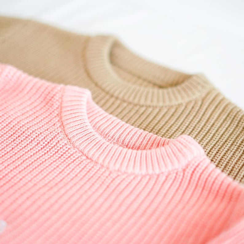 Puede incluir: Primer plano de dos su&eacute;teres de punto. El su&eacute;ter delantero es de color rosa suave, y el de atr&aacute;s es marr&oacute;n claro. Ambos su&eacute;teres tienen una textura de punto acanalado y un cuello redondo. La imagen est&aacute; bien iluminada y muestra los detalles de la tela.