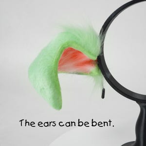 May include: A pair of plush, light green cat ears with a pink interior and white fur accents. The ears are attached to a black headband. Text on the image reads, "The ears can be bent."