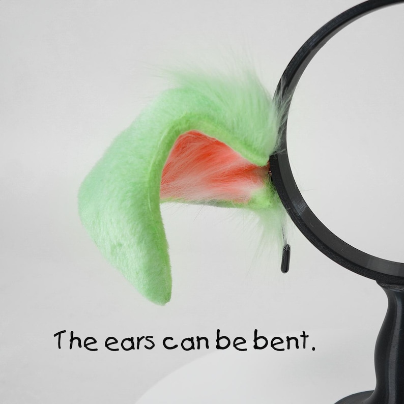 May include: A pair of plush, light green cat ears with a pink interior and white fur accents. The ears are attached to a black headband. Text on the image reads, "The ears can be bent."
