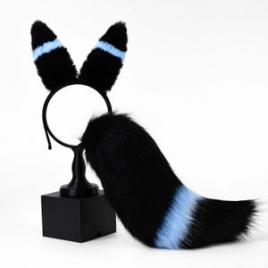 May include: A black faux fur headband featuring two tall, pointed ears with horizontal light blue stripes. A matching long, bushy black faux fur tail is attached, featuring a distinct light blue band near the tip of the tail, creating a set.