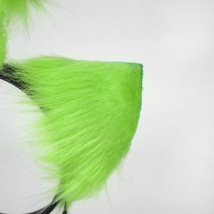 May include: A headband featuring bright neon green faux fur shaped into pointed ears. The long, soft synthetic fibers cover the triangular ear structures, which are attached to a thin, dark-colored plastic band base for secure placement.