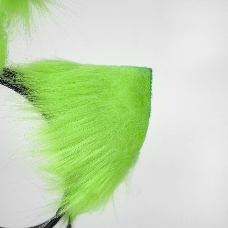 May include: A headband featuring bright neon green faux fur shaped into pointed ears. The long, soft synthetic fibers cover the triangular ear structures, which are attached to a thin, dark-colored plastic band base for secure placement.