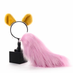 May include: A headband featuring two upright, fuzzy mustard yellow ears attached to a thin black band, paired with a long, flowing, plush pink faux fur tail. The set is displayed on a black stand, showcasing the contrasting textures and colors.