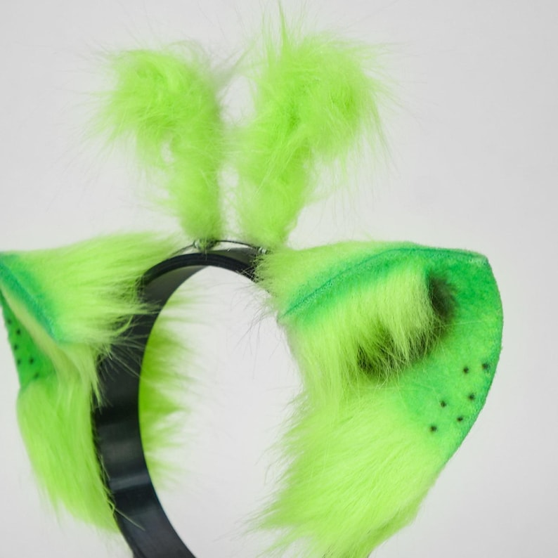May include: A headband featuring bright neon green faux fur ears and two upright tufts of matching green fur on top. The base of the headband is a smooth black plastic band. The ears are triangular with dark green spots on the lower inner sections.