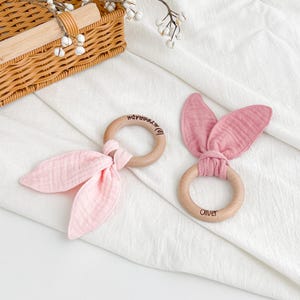May include: Two wooden baby teethers with pink fabric accents. One teether has a bow-tie design, and the other has bunny ear-shaped fabric. The wooden rings have the names "@AUTOGRAPH" and "Oliver" engraved on them. The teethers are on a white surface.