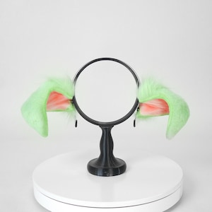 May include: A pair of green and pink faux fur cat ears attached to a black headband. The ears are fluffy and upright, with pink inner ear details. The headband is supported by a black stand on a white surface.