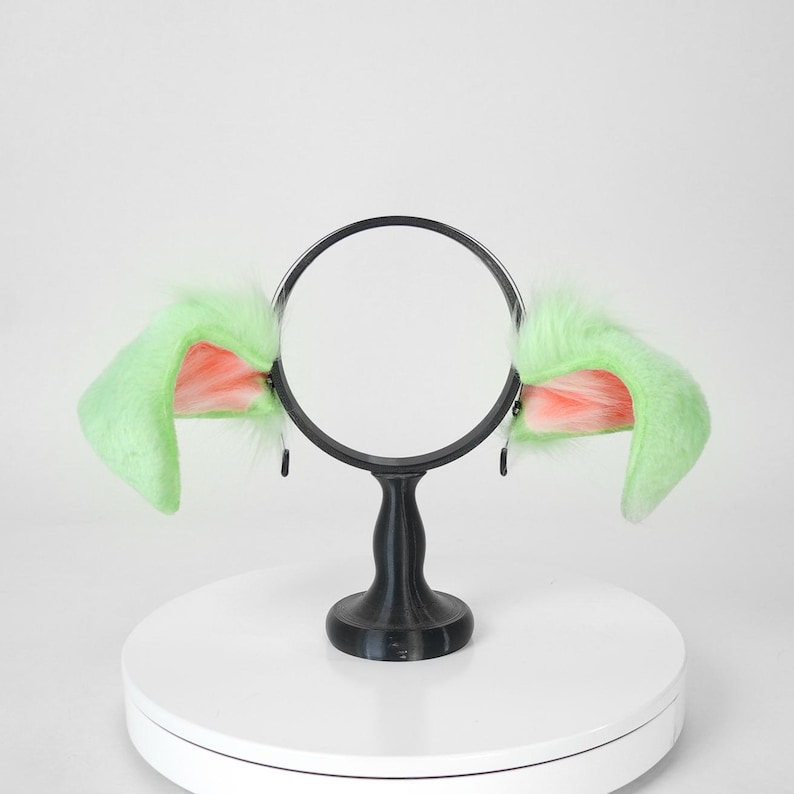 May include: A pair of green and pink faux fur cat ears attached to a black headband. The ears are fluffy and upright, with pink inner ear details. The headband is supported by a black stand on a white surface.