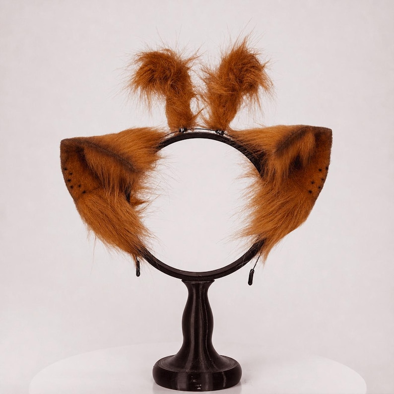 May include: A black headband featuring two large, pointed, brown faux fur animal ears on the sides and two smaller, upright, tufted brown faux fur accents on the top. The ears have dark brown shading and small dot patterns near the outer edges.