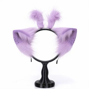May include: A black headband featuring two large, pointed, lavender-colored faux fur animal ears and two smaller, upright, fluffy lavender faux fur tufts on top. The ears have a subtle dotted pattern near the outer edges and long, soft fibers.