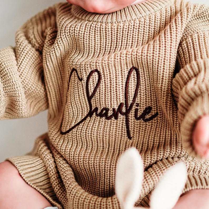 Puede incluir: Un pelele beige para beb&eacute; con el nombre "Charlie" bordado en escritura marr&oacute;n oscuro. El pelele tiene una textura de punto acanalado y mangas largas. Un peluche blanco con orejas largas es parcialmente visible.