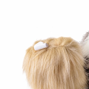 May include: A tan-colored faux fur accessory featuring a soft, shaggy texture and a small, white elastic loop attached to the top edge. The material appears dense and plush, with individual synthetic fibers visible throughout the surface area.