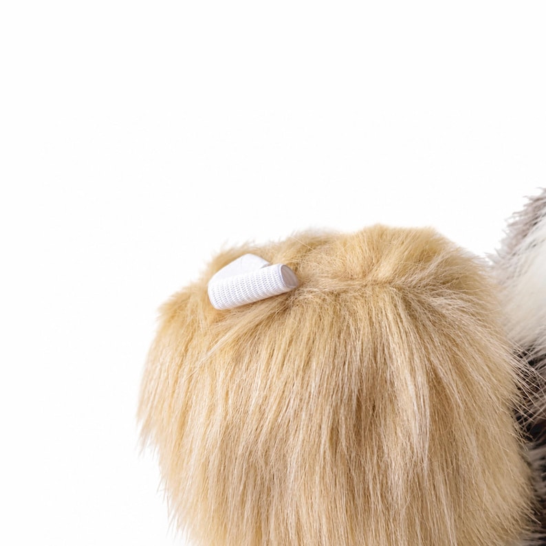 May include: A tan-colored faux fur accessory featuring a soft, shaggy texture and a small, white elastic loop attached to the top edge. The material appears dense and plush, with individual synthetic fibers visible throughout the surface area.
