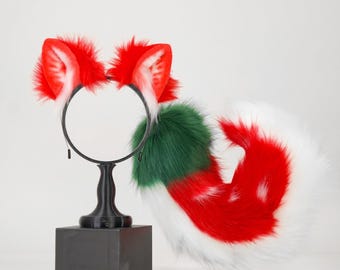 May include: A black headband featuring two pointed, red and white faux fur animal ears, paired with a long, fluffy tail made of green, red, and white faux fur. The set is displayed on a black stand against a plain, light-colored background.