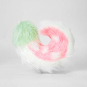 May include: A plush, faux fur tail accessory featuring a curved, multi-colored design. The base is a soft mint green, transitioning into a vibrant pink midsection, and ending in a long, fluffy white tip. The texture is thick and shaggy.