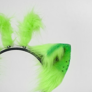 May include: A headband featuring bright lime green faux fur animal ears. The ears are attached to a thin black plastic band with visible silver wire accents. The fur is long and textured, with small dark spots visible on the inner ear surface.