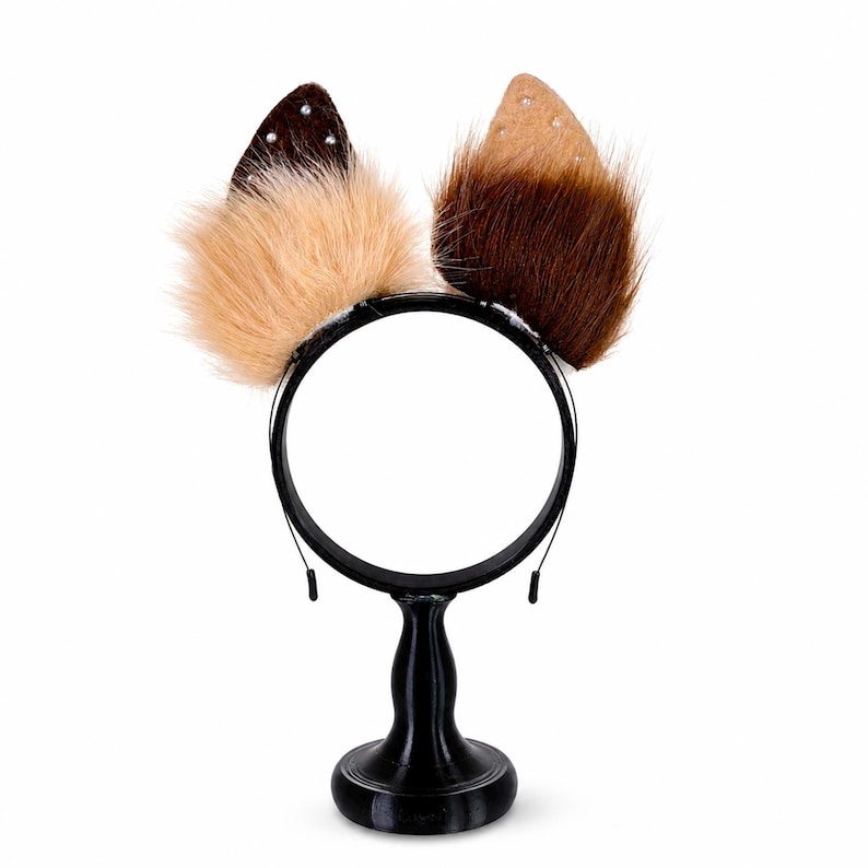 May include: A headband featuring two pointed animal ears. The left ear is dark brown with small white studs and light tan faux fur at the base. The right ear is tan with small white studs and dark brown faux fur at the base. The band is black.