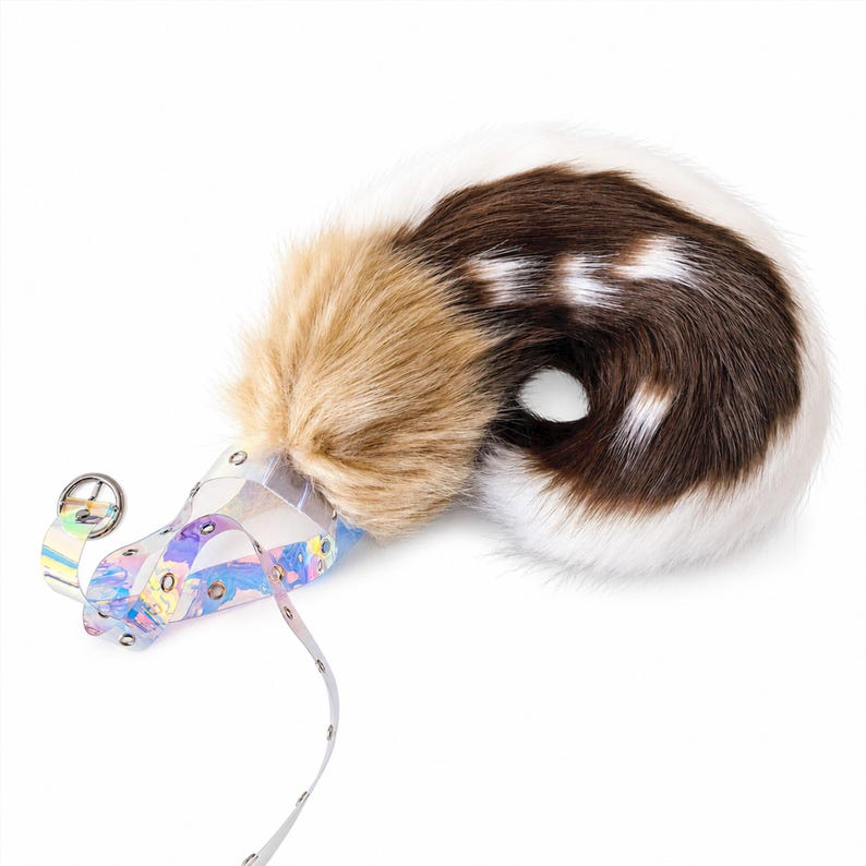 May include: A faux fur animal tail accessory featuring a blend of tan, dark brown, and white fur with a curled shape. The base is attached to a transparent, iridescent plastic harness with silver-toned metal eyelets and a circular buckle.