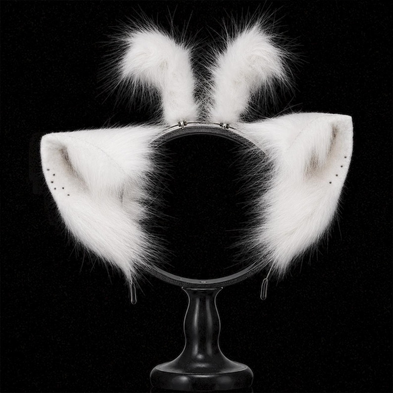 May include: A headband featuring two upright white faux fur ears and two larger, wider white faux fur ears extending to the sides. The fur is long and soft, with small dark dots visible on the inner edges of the side ears. The base is a thin black band.
