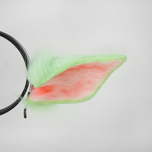 May include: A pair of plush, light green and pink cat ears attached to a black headband. The ears have a fluffy texture and a contrasting pink interior. The headband is black and circular.