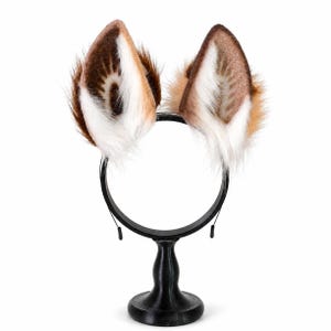 May include: A headband featuring two upright, pointed animal ears made of faux fur. The ears are colored in shades of brown, tan, and white, with long, fluffy white fur at the base and inner sections, and shorter, darker brown fur on the outer edges.