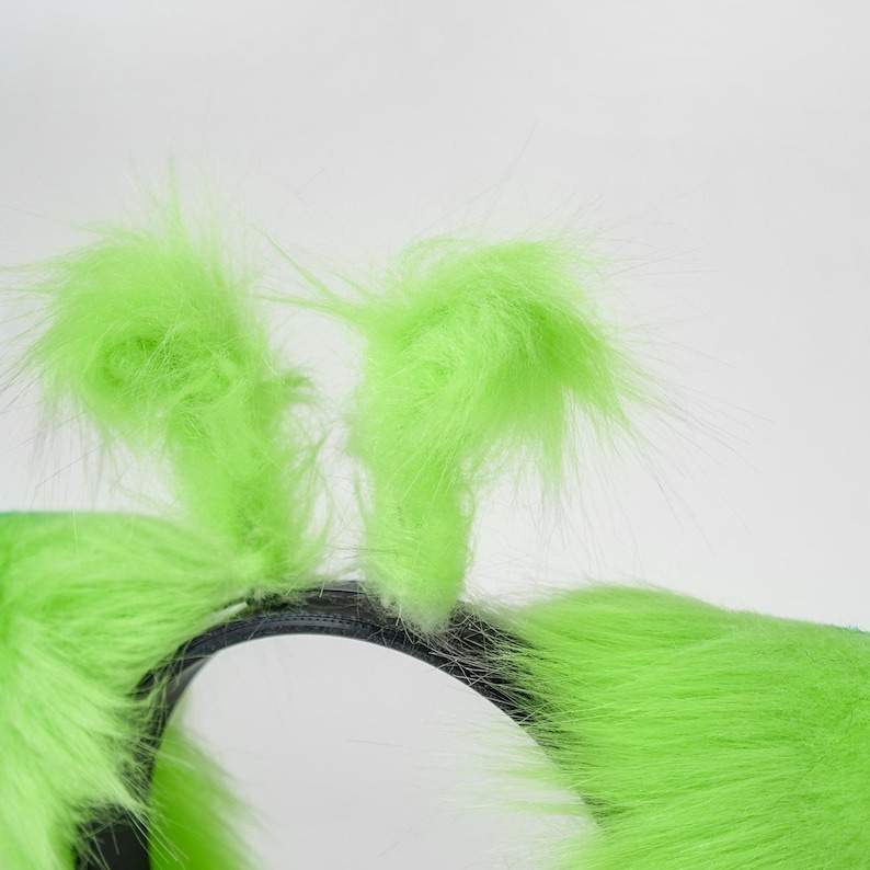 May include: A bright lime green headband featuring two upright, fuzzy, faux fur antennae. The headband base is a thin, black, semi-circular band that supports the two vertical, soft, textured green protrusions extending upward from the top.