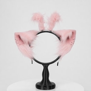 May include: A headband featuring two large, pointed, light pink faux fur ears with small dark spots on the inner edges and two upright, fluffy pink tufts positioned between the ears, all mounted on a thin, black circular metal frame.