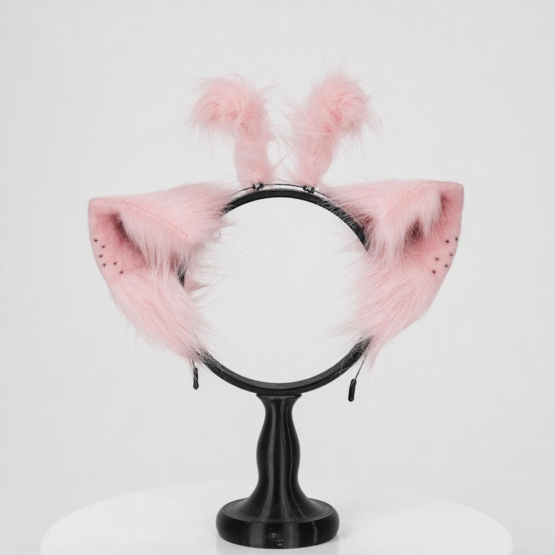 May include: A headband featuring two large, pointed, light pink faux fur ears with small dark spots on the inner edges and two upright, fluffy pink tufts positioned between the ears, all mounted on a thin, black circular metal frame.
